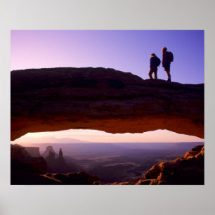 A couple watches sunrise from atop Mesa Arch in Poster