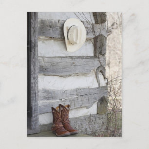 Cowboy boots and hat outside of log cabin postcard