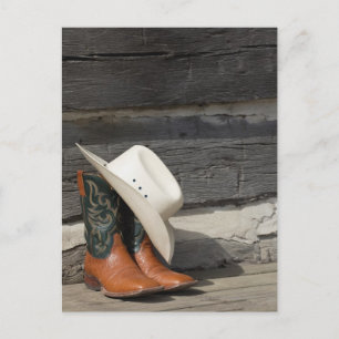Cowboy hat on cowboy boots outside a log cabin postcard