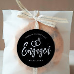 Engagement Party Script Lettering | Black |  Classic Round Sticker<br><div class="desc">A fun and unique white against black script-lettered sticker explicitly tailored to you for an engagement party.  It features the word "engaged" with wedding rings resting over it. Underneath this and above it are the names of the bride and groom and the engagement date.</div>