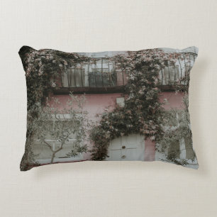 PINK AND WHITE PAINTED HOUSE COVERED WITH PLANTS A DECORATIVE CUSHION
