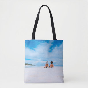 Two women lying on white sand facing beach under b tote bag
