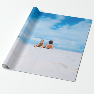 Two women lying on white sand facing beach under b wrapping paper