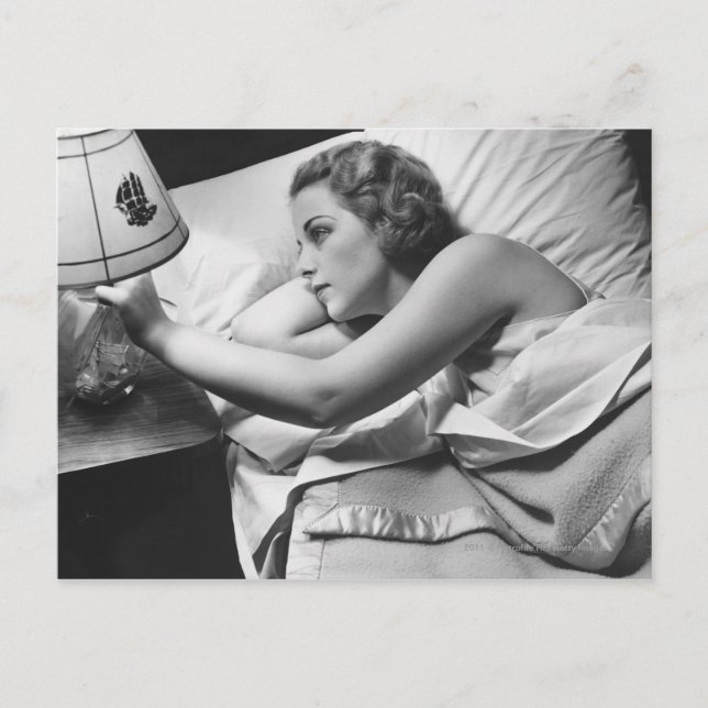 Young woman lying on bed turning off lamp on postcard (Front)